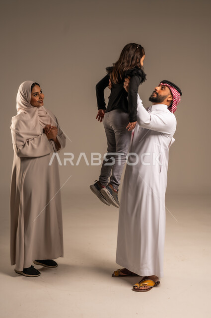 Feelings of love and affection between parents and children, playing, entertainment and fun, portrait of a Saudi Arabian Gulf family having a good time, a Saudi man wearing traditional thobe and shemagh raising his son high with gestures of happiness and pleasure, concept of family stability, gray background