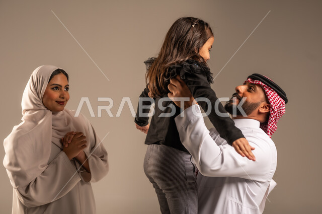 مشاعر الحب والعطف بين الآباء والأبناء، اللعب والتسلية والترفيه، بورتريه لعائلة عربية خليجية سعودية تقضي أوقات ممتعه، رجل سعودي يرتدي الثوب التقليدي والشماغ يرفع ابنه عاليا بإيماءات  السعادة والسرور، مفهوم الاستقرار العائلي, خلفية رمادية