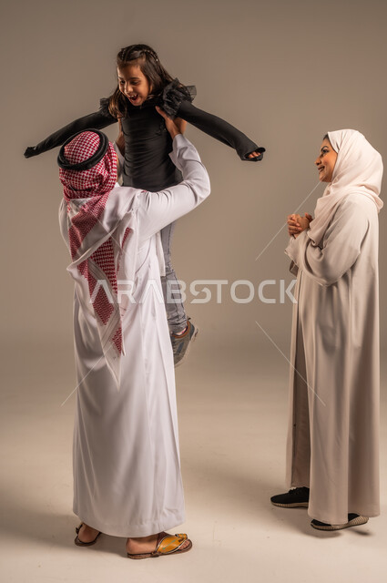 مشاعر الحب والعطف بين الآباء والأبناء، اللعب والتسلية والترفيه، بورتريه لعائلة عربية خليجية سعودية تقضي أوقات ممتعه، رجل سعودي يرتدي الثوب التقليدي والشماغ يرفع ابنه عاليا بإيماءات  السعادة والسرور، مفهوم الاستقرار العائلي, خلفية رمادية