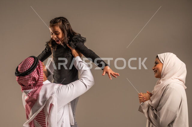 مشاعر الحب والعطف بين الآباء والأبناء، اللعب والتسلية والترفيه، بورتريه لعائلة عربية خليجية سعودية تقضي أوقات ممتعه، رجل سعودي يرتدي الثوب التقليدي والشماغ يرفع ابنه عاليا بإيماءات  السعادة والسرور، مفهوم الاستقرار العائلي, خلفية رمادية
