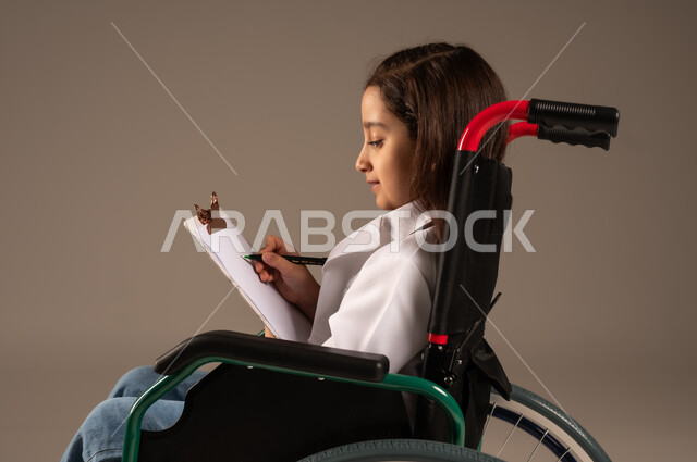 الطموحات و الأهداف المستقبلية في العمل في العيادات والمشافي، تدوين وتسجيل معلومات وبيانات المرضى، تحديد مهنة المستقبل منذ الصغر، بورتريه لفتاة عربية خليجية سعودية من ذوي الاحتياجات الخاصة ترتدي زي ممرضة تجلس على كرسي متحرك تمسك بيدها ملف ملاحظات، خلفية رمادية