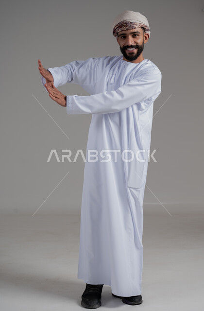 الاحتياط والابتعاد عن شيء ما, حماية النفس من ضرر خارجي, رفع اليدين بإيماءات التوقف وعدم الاقتراب,  بورتريه لرجل عربي خليجي عماني يرتدي الدشداشة والعمامة ينظر الى الكاميرا بتعابير السعادة والسرور, الجسم بالطول الكامل, خلفية رمادي