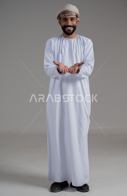 الوقوف باستقامة والنظر إلى الكاميرا بإيماءات السرور, بورتريه لرجل عربي خليجي عماني مبتسم يرتدي الدشداشة والعمامة يفتح كفيه للأعلى بإيماءات رفع عنصر ما, صورة الجسم بالطول الكامل, خلفية رمادي