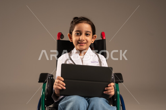 Improving the lives of people with motor disabilities, future ambitions and goals in working in clinics and hospitals, using modern technical devices, portrait of a Saudi Arabian Gulf girl with special needs wearing a nurse's uniform sitting in a wheelchair holding a tablet in her hand, gray background