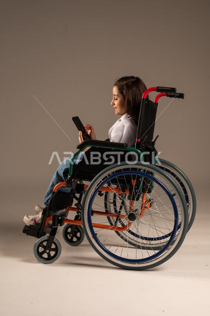الدردشة والتحدث مع الاصدقاء عبر الانترنت، الطموحات والأهداف المستقبلية في العمل في العيادات والمشافي، استخدام الأجهزة التقنية الحديثة، بورتريه لفتاة عربية خليجية سعودية ترتدي زي ممرضة تجلس على كرسي متحرك تمسك بيدها الهاتف المحمول، خلفية رمادية