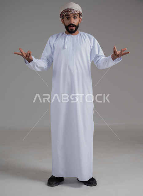 الشعور بالحيرة وعدم المعرفة، بورتريه لرجل عربي عماني خليجي يرتدي الدشداشة والعمامة يفتح كفيه بإيماءات عدم التأكد, التردد وعدم الثقة بخصوص أمر معين، الوقوف باستقامة والنظر إلى الكاميرا، صورة الجسم بالطول الكامل,خلفية رمادي