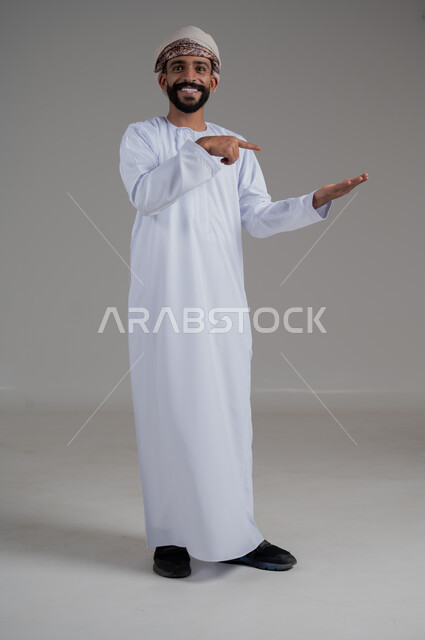 النظر إلى الكاميرا بإيماءات السرور، رفع وحمل شيء ما، بورتريه لرجل عربي خليجي عماني مبتسم يرتدي الدشداشة والعمامة يشير باصبع السبابة على يده، تعابير حمل عنصر، صورة الجسم بالطول الكامل، خلفية رمادي