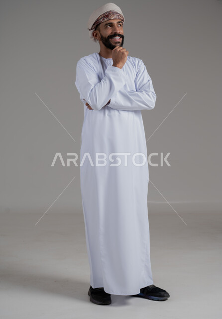 التأمل والتخطيط والتمعن بأمر محدد, بورتريه لرجل عربي خليجي عماني يرتدي الدشداشة والعمامة يضع يديه على ذقنه بإيماءات التفكير, ايجاد الافكار والحلول, صورة الجسم بالطول الكامل, خلفية رمادي