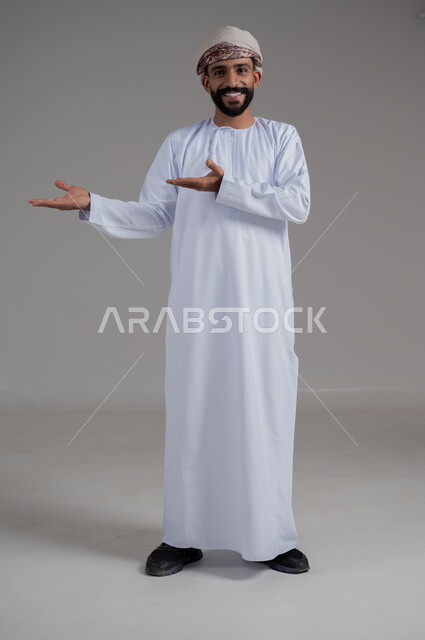 الاهتمام بالمظهر الخارجي, النظر إلى الكاميرا بتعابير السرور, بورتريه لرجل مبتسم عربي خليجي عماني يرتدي الدشداشة و العمامة ينظر إلى الكاميرا و يشير بيديه إلى شيء ما, صورة للجسم بالطول الكامل, خلفية رمادي