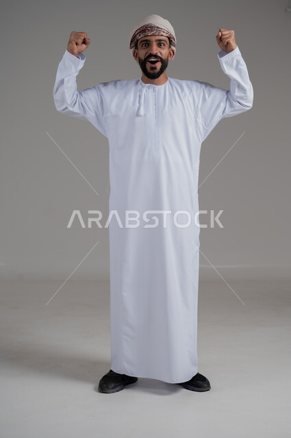 النجاح والقوة وتحقيق الاهداف، بورتريه لرجل عربي خليجي عماني يرتدي العمامة والدشداشة، إيماءات يدين تدل على الحماس والشغف، المنافسة والتحدي والتشجيع، الفرح والسعادة بالفوز والإنتصار، صورة الجسم بالطول الكامل, خلفية رمادي