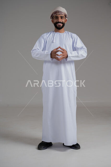 النظر الى الكاميرا يايماءات الثقة بالنفس، تعابير السعادة والسرور، بورتريه لرجل عربي خليجي عماني يرتدي الدشداشة والعمامة يرفع يديه بحركة تلامس الأصابع، مفهوم الذكاء والمعرفة، صورة الجسم بالطول الكامل، خلفية رمادي