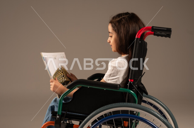 تنمية المهارات الفنية والذهنية، استغلال أوقات الفراغ بالأنشطة التعليمية، بورتريه مقرب من الجانب لفتاة عربية خليجية سعودية من ذوي الاحتياجات الخاصة ترتدي الزي الكاجوال تجلس على كرسي متحرك تقوم بالمطالعة وقراءة الكتب المفيده، مراجعة الدروس اليومية، خلفية رمادية