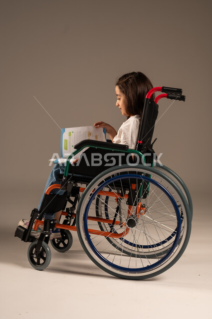 تنمية المهارات الفنية والذهنية، استغلال أوقات الفراغ بالأنشطة التعليمية، بورتريه من الجانب لفتاة عربية خليجية سعودية من ذوي الاحتياجات الخاصة ترتدي الزي الكاجوال تجلس على كرسي متحرك تقوم بالمطالعة وقراءة الكتب المفيده، مراجعة الدروس اليومية، خلفية رمادية