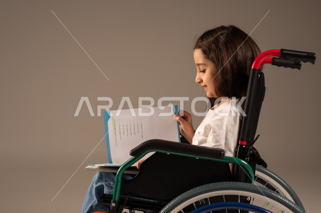 تنمية المهارات الفنية والذهنية، استغلال أوقات الفراغ بالأنشطة التعليمية، بورتريه من الجانب لفتاة عربية خليجية سعودية من ذوي الاحتياجات الخاصة ترتدي الزي الكاجوال تجلس على كرسي متحرك تقوم بالمطالعة وقراءة الكتب المفيده، مراجعة الدروس اليومية، خلفية رمادية