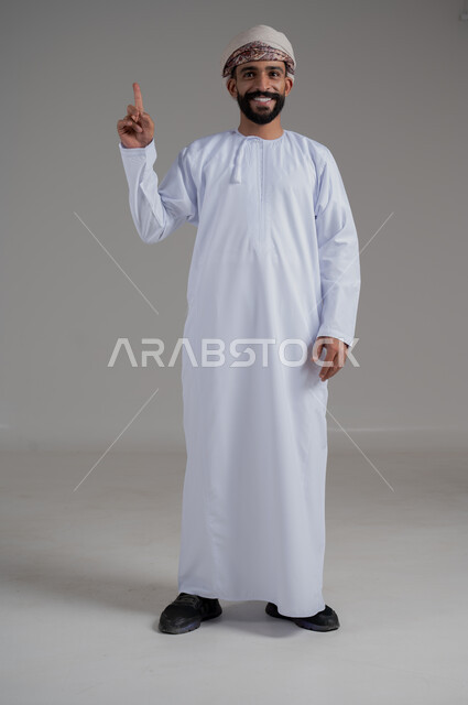 الاهتمام بالمظهر الخارجي, النظر إلى الكاميرا بتعابير السرور, بورتريه لرجل مبتسم عربي خليجي عماني يرتدي الدشداشة و العمامة ينظر إلى الكاميرا و يشير باصبع السبابة إلى الاعلى, صورة للجسم بالطول الكامل, خلفية رمادي
