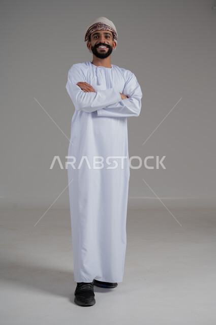 الاهتمام بالمظهر الخارجي, النظر إلى الكاميرا بتعابير السرور, بورتريه لرجل مبتسم عربي خليجي عماني يرتدي الدشداشة و العمامة ينظر إلى الكاميرا ويقف بايدي متقاطعة, صورة للجسم بالطول الكامل, خلفية رمادي