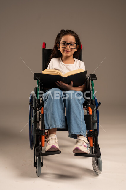 مراجعة الدروس اليومية، استغلال أوقات الفراغ بالأنشطة التعليمية، تنمية المهارات الفنية والذهنية، بورتريه لفتاة عربية خليجية سعودية من ذوي الاحتياجات الخاصة ترتدي الزي الكاجوال تجلس على كرسي متحرك تقوم بالمطالعة وقراءة الكتب المفيده، خلفية رمادية