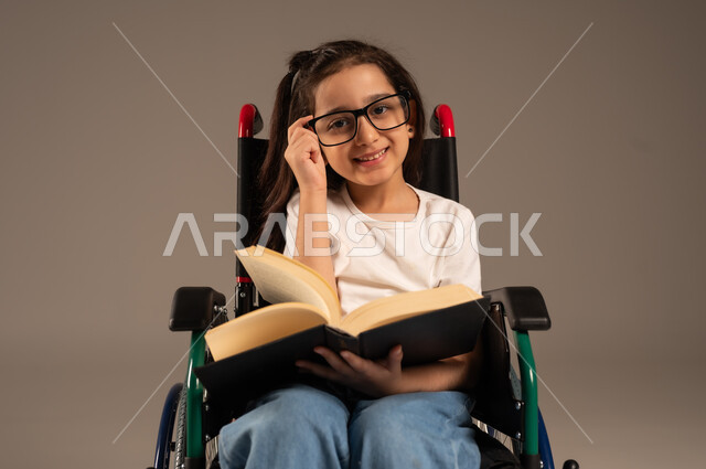مراجعة الدروس اليومية، استغلال أوقات الفراغ بالأنشطة التعليمية، تنمية المهارات الفنية والذهنية، بورتريه لفتاة عربية خليجية سعودية من ذوي الاحتياجات الخاصة ترتدي الزي الكاجوال تجلس على كرسي متحرك تقوم بالمطالعة وقراءة الكتب المفيده، خلفية رمادية