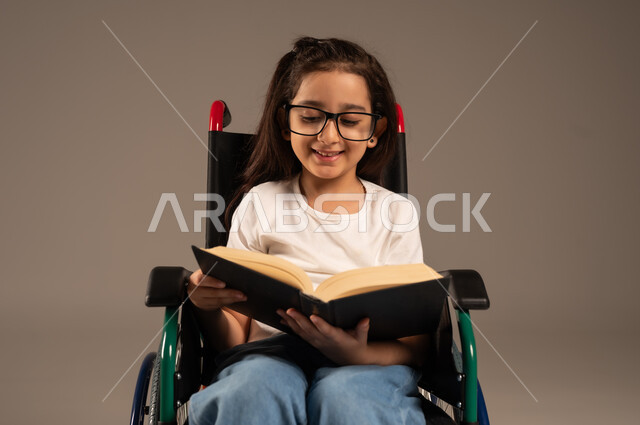 مراجعة الدروس اليومية، استغلال أوقات الفراغ بالأنشطة التعليمية، تنمية المهارات الفنية والذهنية، بورتريه لفتاة عربية خليجية سعودية من ذوي الاحتياجات الخاصة ترتدي الزي الكاجوال تجلس على كرسي متحرك تقوم بالمطالعة وقراءة الكتب المفيده، خلفية رمادية