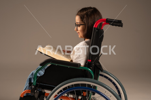 مراجعة الدروس اليومية، استغلال أوقات الفراغ بالأنشطة التعليمية، تنمية المهارات الفنية والذهنية، بورتريه من الجانب لفتاة عربية خليجية سعودية من ذوي الاحتياجات الخاصة ترتدي الزي الكاجوال تجلس على كرسي متحرك تقوم بالمطالعة وقراءة الكتب المفيده، خلفية رمادية