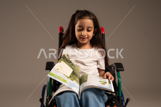  تنمية المهارات الفنية والذهنية، مراجعة الدروس اليومية، استغلال أوقات الفراغ بالأنشطة التعليمية، بورتريه لفتاة عربية خليجية سعودية من ذوي الاحتياجات الخاصة ترتدي الزي الكاجوال تجلس على كرسي متحرك تقوم بالمطالعة وقراءة الكتب المفيده، خلفية رمادية