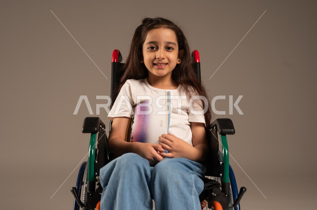 Developing technical and mental skills, reviewing daily lessons, exploiting free time with educational activities, portrait of a Saudi Arabian Gulf girl with special needs wearing casual clothes sitting in a wheelchair reading useful books, gray background