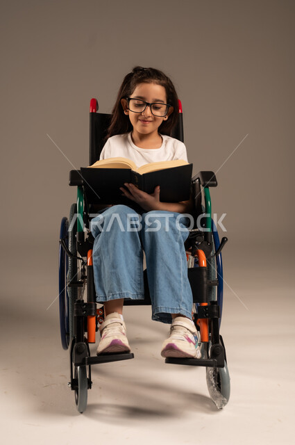 Developing technical and mental skills, reviewing daily lessons, exploiting free time with educational activities, portrait of a Saudi Arabian Gulf girl with special needs wearing casual clothes sitting in a wheelchair reading useful books, gray background