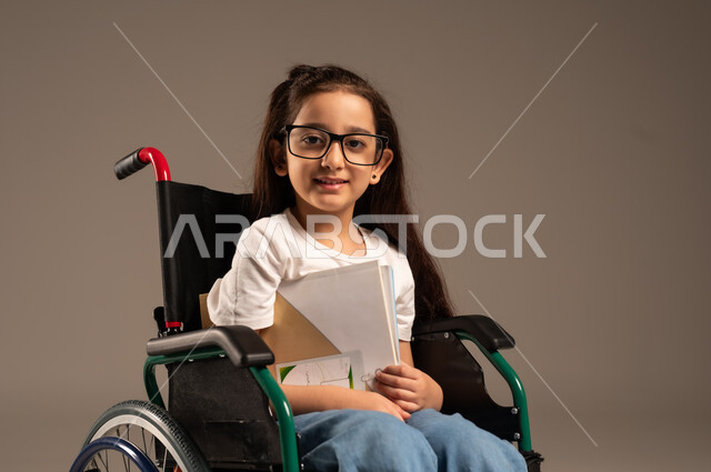 Utilizing free time in educational activities, portrait of a Saudi Arabian Gulf girl with special needs wearing casual clothes sitting in a wheelchair reading useful books, developing artistic and mental skills, reviewing daily lessons, gray background