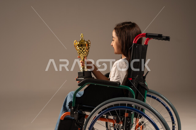 التتويج بالبطولة بعد الفوز والحصول على المركز الأول، بورتريه من الجانب لفتاة عربية خليجية سعودية ترتدي الزي الكاجوال تجلس على كرسي متحرك تحمل بيدها الكأس الذهبي بحماس، تحسين حياة ذوي الإعاقة الحركية، مفهوم الربح والفوز، خلفية رمادية