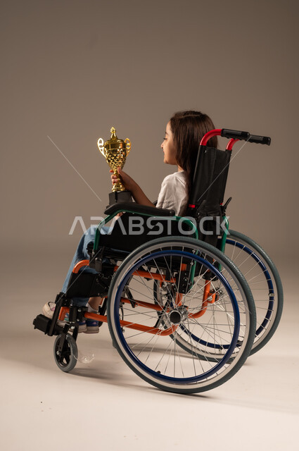 Crowning the championship after winning and getting first place, portrait of a Saudi Arabian Gulf girl wearing casual clothes sitting on a wheelchair holding the golden cup in her hand with enthusiasm, improving the lives of people with motor disabilities, the concept of profit and victory, gray background