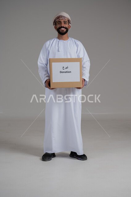 مفهوم التبرع والأعمال الخيرية الإسلاميه في سلطنه عمان، بورتريه لرجل عربي عماني خليجي يرتدي الدشداشة والعمامة يمسك بيده صندوق كرتوني، النظر إلى الكاميرا بإيماءات السرور، خلفية رمادي
