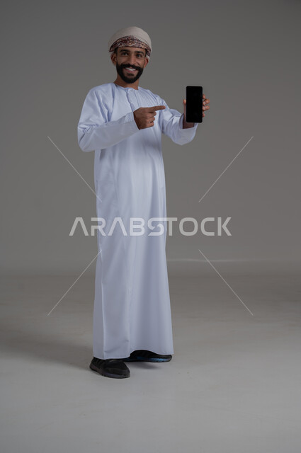 استخدام التكنولوجيا و التقنيات الحديثة المتطورة، بورتريه لرجل عربي خليجي عماني يرتدي الدشداشه والعمامة يقوم بعرض شاشة سوداء فارغة عبر الهاتف المحمول ويشير باصبعه السبابة نحوها، النظر الى الكاميرا بتعابير السعادة والسرور، صورة الجسم بالطول الكامل خلفية رمادي