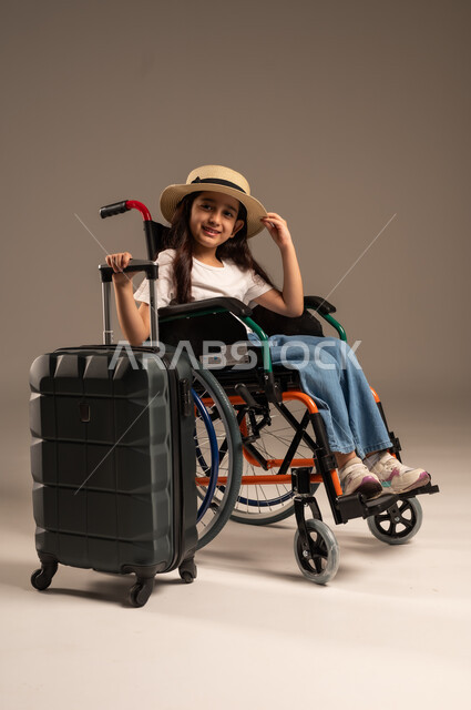 القيام بالجولات الترفيهية وقت الإجازة، الاستعداد لقضاء إجازة صيفية ممتعة, بورتريه لفتاة عربية خليجية سعودية من ذوي الهمم ترتدي الزي الكاجوال تجلس على كرسي متحرك وبجانبها حقيبة سفر سوادء، تأمين اجهزة ومعدات تساعد بالحركة والتنقل، خلفية رمادية