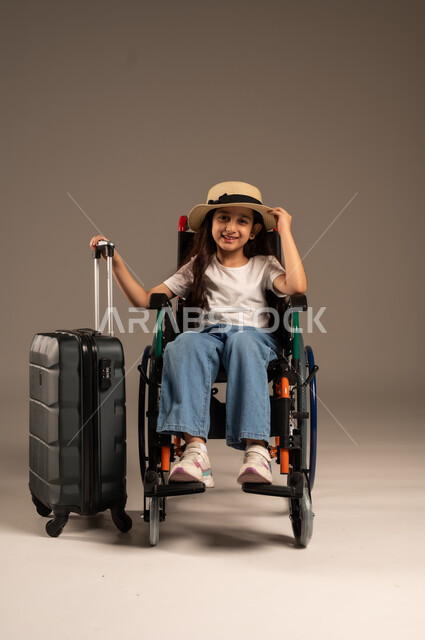 القيام بالجولات الترفيهية وقت الإجازة، الاستعداد لقضاء إجازة صيفية ممتعة, بورتريه لفتاة عربية خليجية سعودية من ذوي الهمم ترتدي الزي الكاجوال تجلس على كرسي متحرك وبجانبها حقيبة سفر سوادء، تأمين اجهزة ومعدات تساعد بالحركة والتنقل، خلفية رمادية