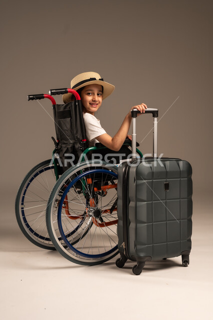 القيام بالجولات الترفيهية وقت الإجازة، الاستعداد لقضاء إجازة صيفية ممتعة, بورتريه من الجانب لفتاة عربية خليجية سعودية من ذوي الهمم ترتدي الزي الكاجوال تجلس على كرسي متحرك وبجانبها حقيبة سفر سوادء، تأمين اجهزة ومعدات تساعد بالحركة والتنقل، خلفية رمادية