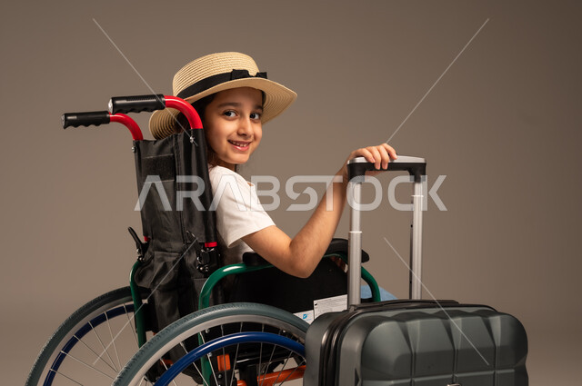 القيام بالجولات الترفيهية وقت الإجازة، الاستعداد لقضاء إجازة صيفية ممتعة, بورتريه مقرب من الجانب لفتاة عربية خليجية سعودية من ذوي الهمم ترتدي الزي الكاجوال تجلس على كرسي متحرك وبجانبها حقيبة سفر سوادء، تأمين اجهزة ومعدات تساعد بالحركة والتنقل، خلفية رمادية