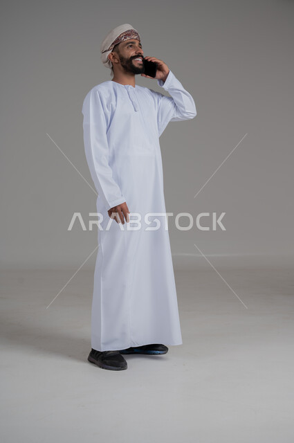 الدردشة مع الأهل والأصدقاء، تسهيل التواصل مع الآخرين باستخدام الهاتف المحمول، تطورات تقنية وتكنولوجية، بورتريه لرجل عماني يرتدي الدشداشة والعمامة يقوم بالمحادثة مع الأقارب عبر الموبايل، صورة الجسم بالطول الكامل, خلفية رمادي