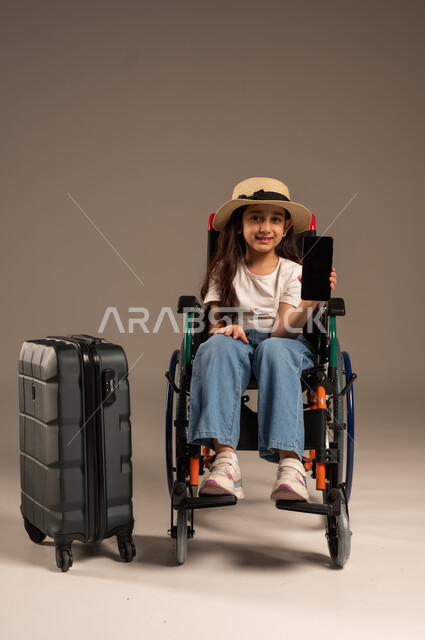 عرض شاشة سوداء فارغة عبر الهاتف المحمول، الاستعداد لقضاء إجازة صيفية ممتعة, بورتريه مقرب لفتاة عربية خليجية سعودية من ذوي الهمم ترتدي الزي الكاجوال تجلس على كرسي متحرك وبجانبها حقيبة سفر سوادء، تأمين اجهزة ومعدات تساعد بالحركة والتنقل، خلفية رمادية