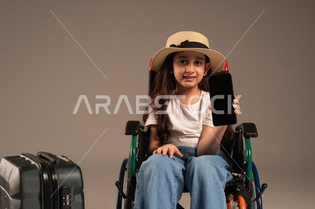 عرض شاشة سوداء فارغة عبر الهاتف المحمول، الاستعداد لقضاء إجازة صيفية ممتعة, بورتريه لفتاة عربية خليجية سعودية من ذوي الهمم ترتدي الزي الكاجوال تجلس على كرسي متحرك وبجانبها حقيبة سفر سوادء، تأمين اجهزة ومعدات تساعد بالحركة والتنقل، خلفية رمادية