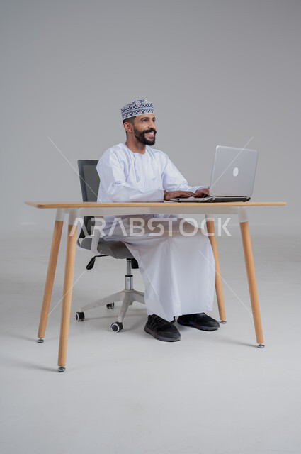 مهن ووظائف مكتبيه عمانية، استخدام التقنيات الحديثة والمتطورة، بورتريه لرجل عربي خليجي عماني مبتسم يترتدي الدشداشة والكمة يجلس على الكرسي أمام الحاسوب المحمول,  خلفية رمادي