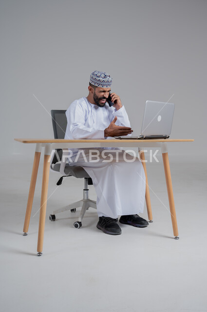 ايماءات الغضب والانزعاج, تعابير تلقي خبر مزعج عبر الهاتف المحمول, مهن ووظائف مكتبيه عمانية، استخدام التقنيات الحديثة والمتطورة، بورتريه لرجل عربي خليجي عماني غاضب يترتدي الدشداشة والكمة يجلس على الكرسي أمام الحاسوب المحمول,  خلفية رمادي