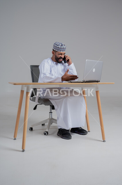 ايماءات الغضب والانزعاج, تعابير تلقي خبر مزعج عبر الهاتف المحمول, مهن ووظائف مكتبيه عمانية، استخدام التقنيات الحديثة والمتطورة، بورتريه لرجل عربي خليجي عماني غاضب يترتدي الدشداشة والكمة يجلس على الكرسي أمام الحاسوب المحمول,  خلفية رمادي
