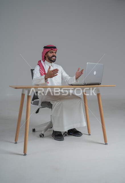 الحماس والقيام بصفقات ناجحة مع العملاء عبر الحاسب المحمول, بورتريه لرجل عربي خليجي سعودي يرتدي الثوب التقليدي والشماغ يجلس على كرسي وامامه المكتب, تعابير الفوز والنصر وتحقيق أهداف العمل الاداري, ايماءات وجه ويدين تدل على السرور والبشاشة وإنجاز المهام المطلوبة, خلفية رمادي