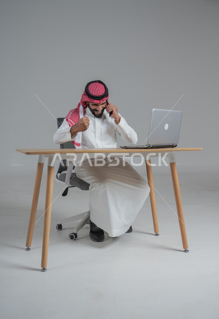 الحماس والقيام بصفقات ناجحة مع العملاء عبر الهاتف المحمول, بورتريه لرجل عربي خليجي سعودي يرتدي الثوب التقليدي والشماغ يجلس على كرسي وامامه المكتب, تعابير الفوز والنصر وتحقيق أهداف العمل الاداري, ايماءات وجه ويدين تدل على السرور والبشاشة وإنجاز المهام المطلوبة, خلفية رمادي