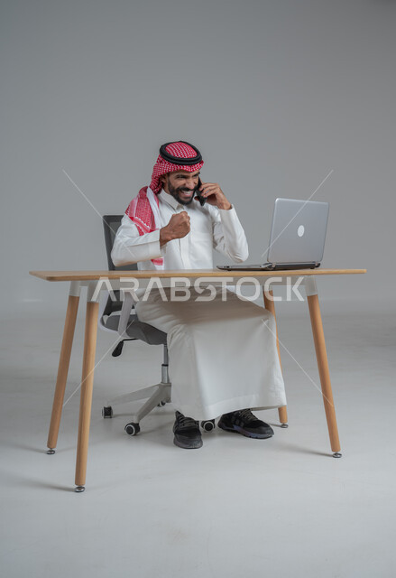الحماس والقيام بصفقات ناجحة مع العملاء عبر الهاتف المحمول, بورتريه لرجل عربي خليجي سعودي يرتدي الثوب التقليدي والشماغ يجلس على كرسي وامامه المكتب, تعابير الفوز والنصر وتحقيق أهداف العمل الاداري, ايماءات وجه ويدين تدل على السرور والبشاشة وإنجاز المهام المطلوبة, خلفية رمادي