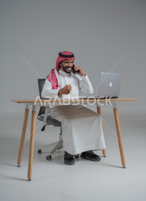 Enthusiasm and making successful deals with customers via mobile phone, portrait of a Saudi Gulf Arab man wearing a traditional thobe and shemagh sitting on a chair with a desk in front of him, expressions of victory and victory and achieving the goals of administrative work, facial and hand gestures that indicate pleasure, cheerfulness and the completion of required tasks, gray background
