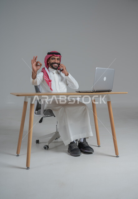 الحماس والقيام بصفقات ناجحة مع العملاء عبر الهاتف المحمول, بورتريه لرجل عربي خليجي سعودي يرتدي الثوب التقليدي والشماغ يجلس على كرسي وامامه المكتب, تعابير الفوز والنصر وتحقيق أهداف العمل الاداري, ايماءات وجه ويدين تدل على السرور والبشاشة وإنجاز المهام المطلوبة, خلفية رمادي