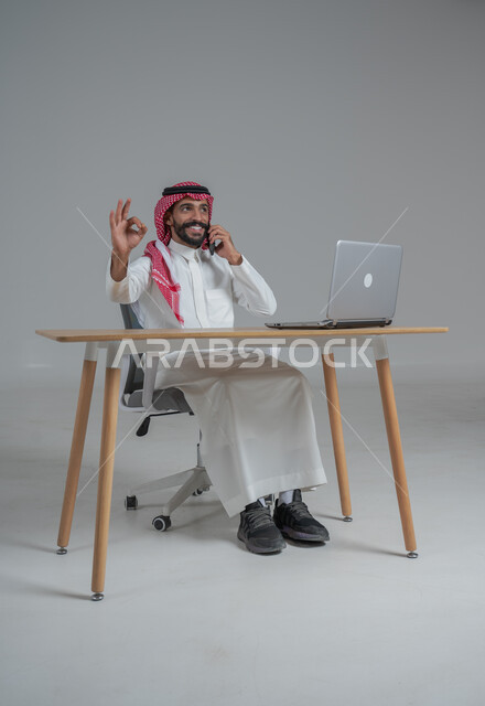 الحماس والقيام بصفقات ناجحة مع العملاء عبر الهاتف المحمول, بورتريه لرجل عربي خليجي سعودي يرتدي الثوب التقليدي والشماغ يجلس على كرسي وامامه المكتب, تعابير الفوز والنصر وتحقيق أهداف العمل الاداري, ايماءات وجه ويدين تدل على السرور والبشاشة وإنجاز المهام المطلوبة, خلفية رمادي