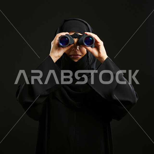 التخطيط المستقبلي للأهداف والطموحات، إمساك المنظار بكلتا اليدين والتركيز على رصد هدف معين، حب الاستكشاف والمغامرة، بورتريه لسيدة عربية خليجية سعودية ترتدي الحجاب والعباءة وتنظر من خلال منظار الرؤية ثنائي العينين، خلفية سوداء