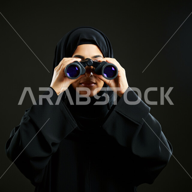 Holding binoculars with both hands and focusing on observing a specific target, love of exploration and adventure, portrait of a Saudi Arabian Gulf woman wearing hijab and abaya and looking through binoculars, future planning for goals and ambitions, black background